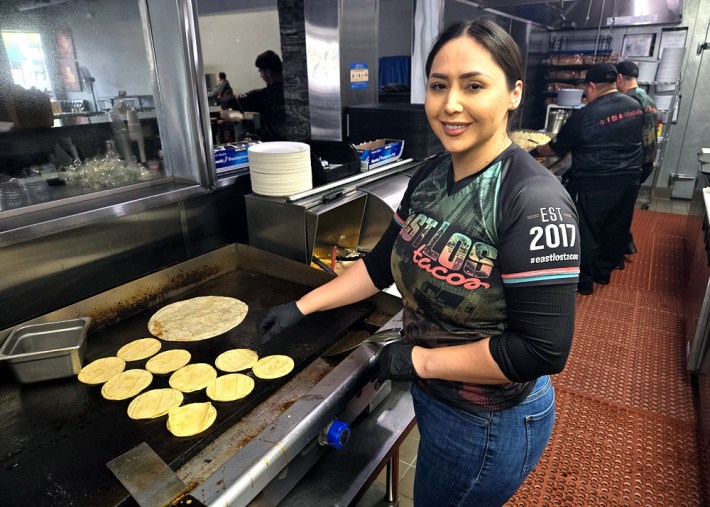 Erika Armenta, owner of East Los Tacos and East Los Pantry, stands over a griddle heating up a flour tortilla and several corn tortillas with her hair pulled back and black gloves and an East Los Tacos shirt on.