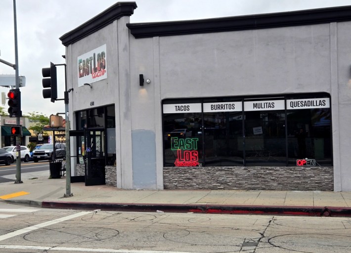 A gray building with a neon sign in the side window that says East Los Tacos. And a white sign on front that says East Los Tacos. Next to a red-lit traffic light.