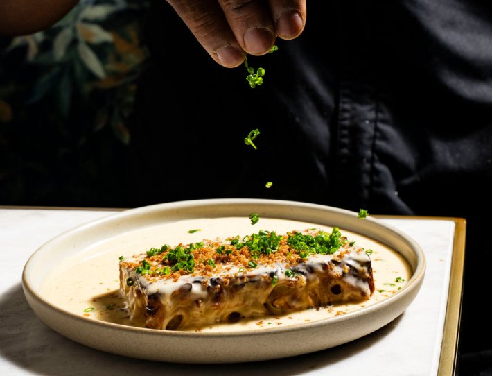 A chef's hand releasing micro herbs onto the top of a Trini mac pie on a plate on a white tabletop.
