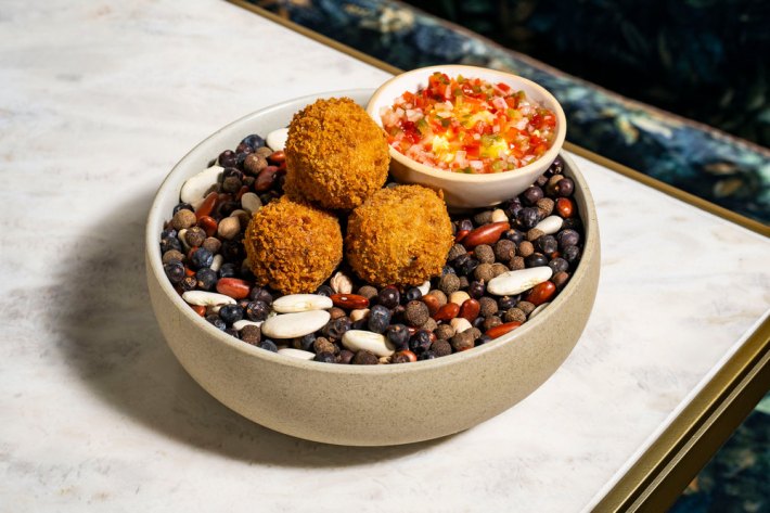 A bowl of uncooked beans supporting three golden croquettes next to a smaller bowl of salsa criolla.