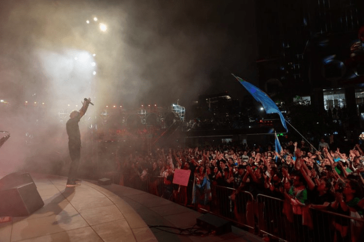 A singer holds his hands above his head before a crowd, some of whom are waving flags