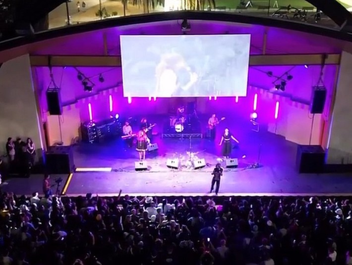 Under purple lighting, a band plays on stage at The Levitt Pavilion, with a screen over their heads and a big crowd in front of them. Photo taken from a drone above.