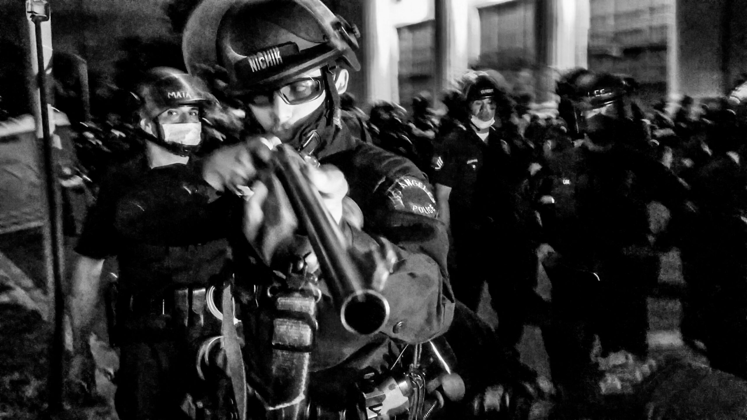 An LAPD officer points a "less-lethal" rifle at a photographer, while a group of officers stands in the background.